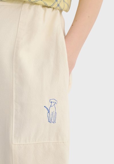 Personne portant un pantalon crème avec un petit chien bleu brodé sur la cuisse et la main placée à l'intérieur de la poche.