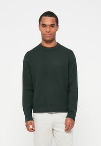 Pulover verde închis cu guler rotund, mâneci lungi și o croială lejeră, realizat dintr-un material tricotat moale și texturat. Asortat cu pantaloni în tonuri deschise.