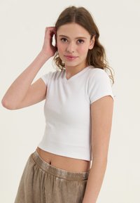 Bílý žebrovaný crop top s krátkými rukávy a výstřihem do V, doplněný o světlé hnědé volné kalhoty s elastickým pasem.