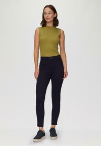 Mouwloze, geribbelde groene top gecombineerd met donker marineblauwe, strakke broek. Model draagt marineblauwe sneakers met een gestructureerde zool. Eenvoudige, moderne silhouet.