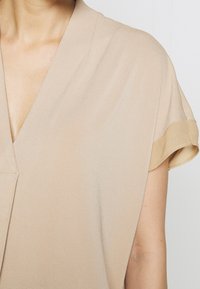 Beige inlindad blus med mjukt draperad tyg, korta rullade ärmar och v-ringning. Sl ät textur utan mönster eller synliga detaljer.