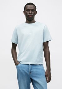 Jasnoniebieska bawełniana koszulka z krótkim rękawem, z subtelnym białym logo na piersi, zestawiona z jasnymi dżinsami w jasnym odcieniu.