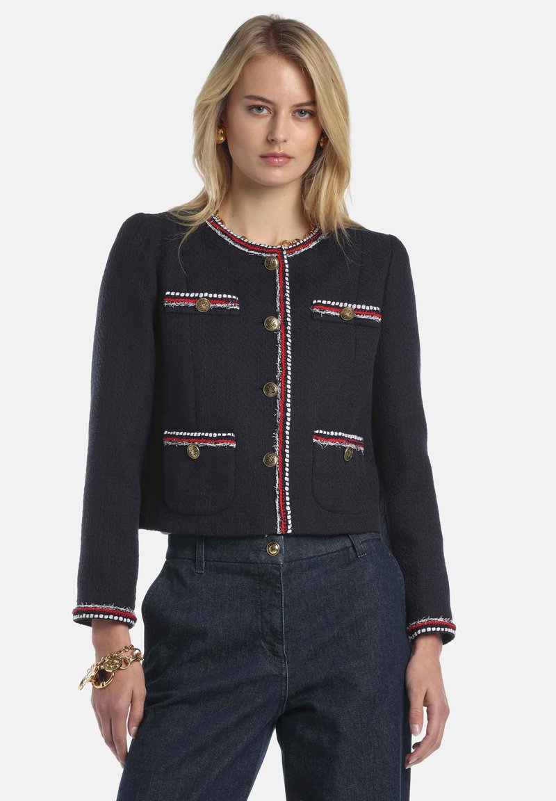 Veste en tissu bleu marine avec boutons dorés, bordures rouges et blanches le long des bords et des poches, présentant un design structuré et une coupe courte.