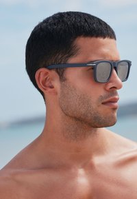 Naočale s mat sivim okvirom, pravokutnim lećama i suptilnim bočnim detaljima. Nosio ih je muški model s kratkom kosom na plaži.