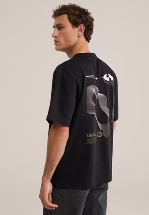 Jeune homme de profil portant un t-shirt noir graphique avec un dessin abstrait en lignes blanches et un petit texte dans le dos, associé à un jean foncé.