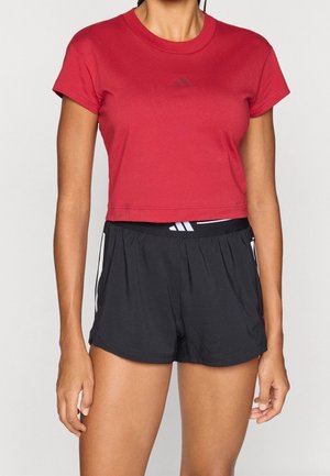 Donna che indossa una maglietta corta rossa e pantaloncini sportivi neri con strisce bianche ai lati su uno sfondo semplice.