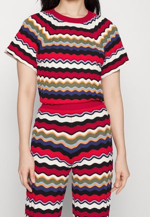 Femme portant un haut à manches courtes et un pantalon assortis avec des rayures ondulées horizontales multicolores en rouge, noir, blanc, bleu, orange et vert.