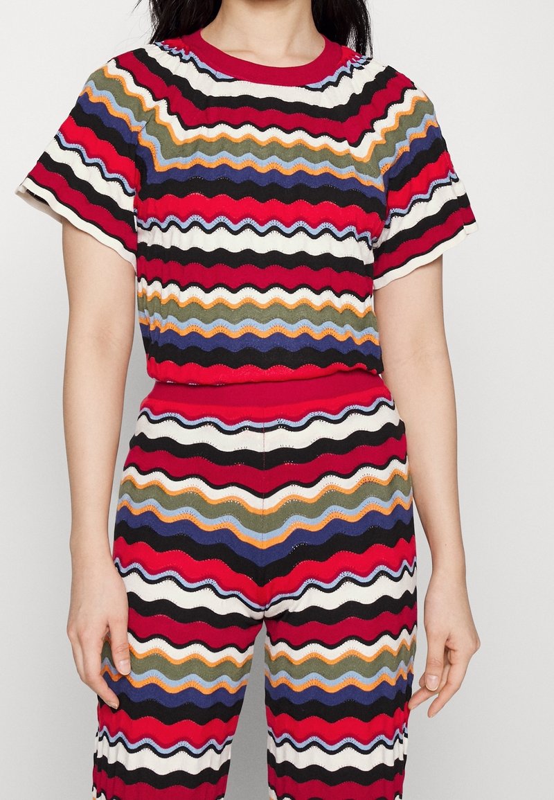 Femme portant un haut à manches courtes et un pantalon assortis avec des rayures ondulées horizontales multicolores en rouge, noir, blanc, bleu, orange et vert.