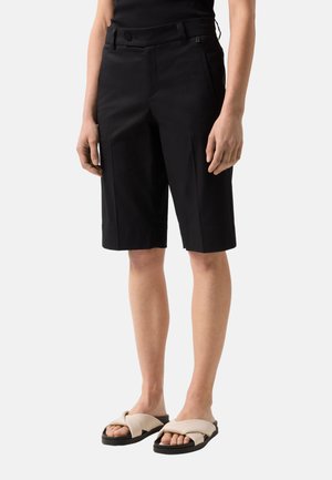 Schwarze, knielange, maßgeschneiderte Shorts, getragen mit beigen Slip-On-Sandalen an einer Person, die vor einem einfarbigen Hintergrund steht.