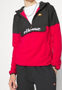 Chaqueta de forro polar roja y negra con media cremallera, que cuenta con un bolsillo delantero y el logo blanco "ellesse". Incluye capucha y puños elásticos.