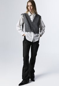 Gilet grigio corto con bordi sfilacciati sopra una camicia bianca, abbinato a pantaloni neri a zampa d'elefante con cuciture bianche a contrasto e tacchi a punta.