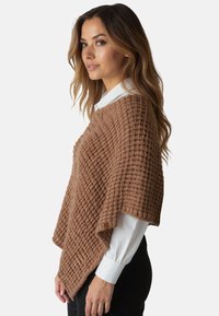 Poncho lavorato a maglia marrone con un motivo a diamante testurizzato che ricade su una camicia bianca con colletto. Il tessuto è morbido e leggero, con un orlo diagonale.