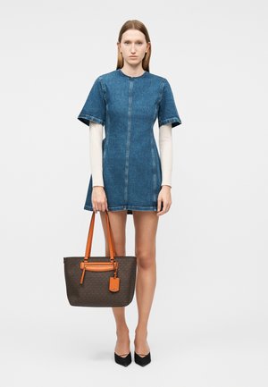 Mujer de pie mirando al frente, lleva un vestido corto de mezclilla azul sobre una camiseta blanca de manga larga, sosteniendo un bolso tote marrón con asas naranjas.