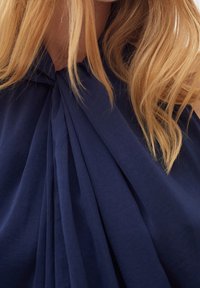 Marinblå veckad tygblus med en knuten design vid halsringningen. Slät, texturerad tyg med flödande draperingar och diskret elegans.