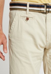 Pantaloni in cotone beige con una finitura testurizzata, dotati di chiusura a bottone e tasche laterali, abbinati a una cintura intrecciata multicolore.