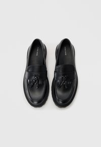 Mocasines de cuero negro con punta redondeada, que cuentan con un detalle decorativo de borlas y una suela texturizada. Superficie superior lisa con acentos de costura.