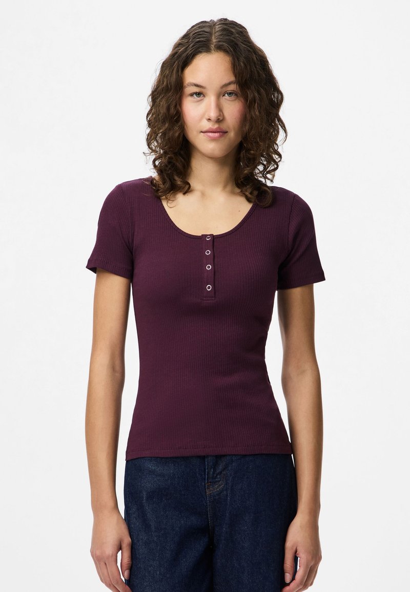 Jauna moteris su garbanotais plaukais, vilkinti prigludusią trumpų rankovių slyvų spalvos šonkaulinę palaidinę su keturiais mygtukais ir tamsiai mėlynas džinsus, stovi prieš baltą foną.