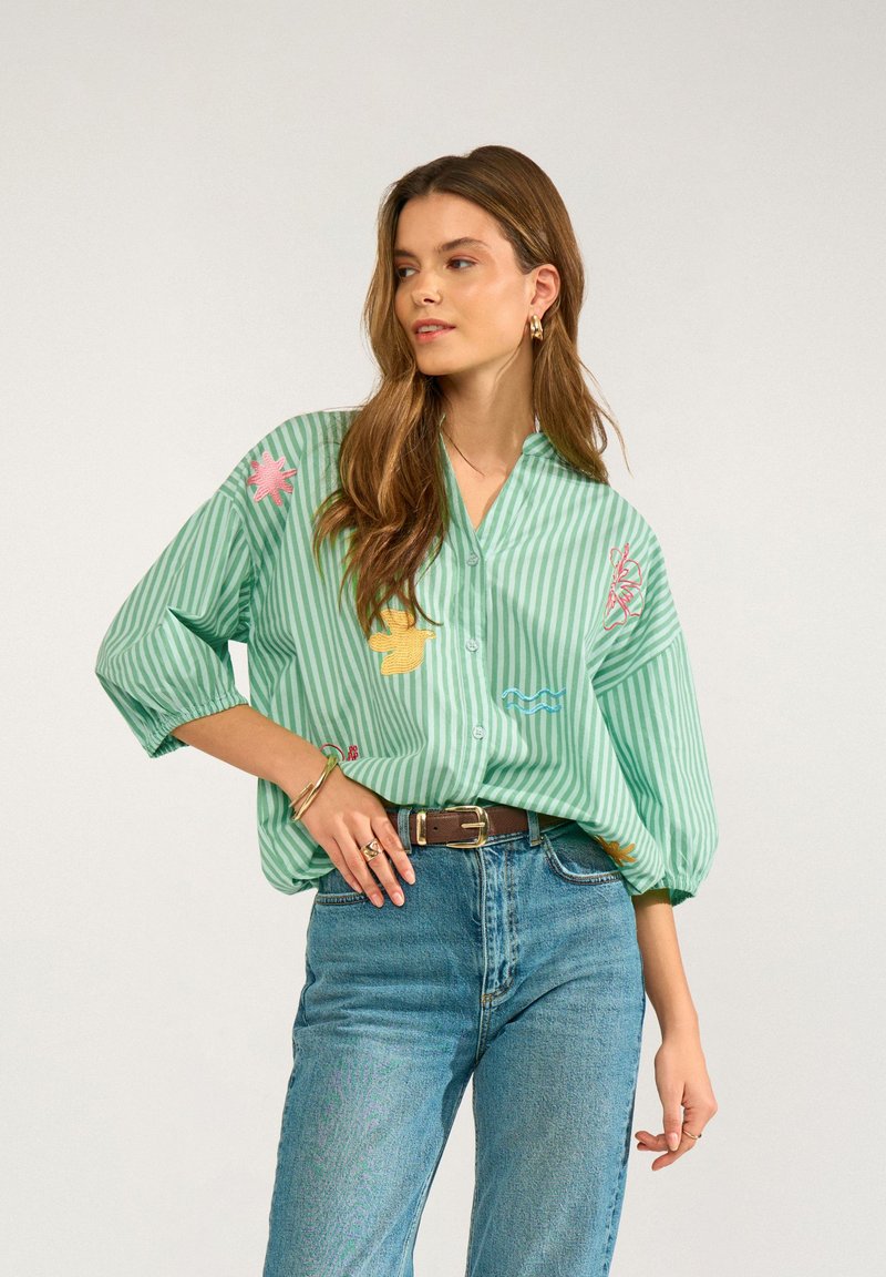 Femme portant une chemise verte à rayures avec une broderie colorée et un jean bleu, posant avec une main sur la hanche devant un fond uni.