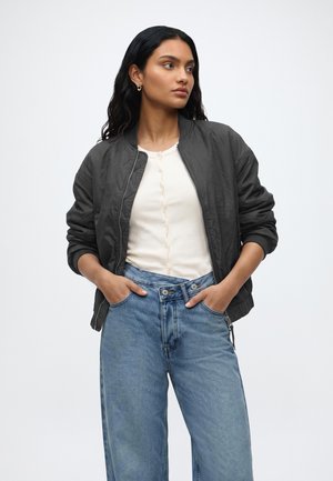 Femme aux longs cheveux foncés portant une veste bomber noire, un haut blanc boutonné et un jean bleu taille haute, les mains dans les poches, regardant sur le côté.