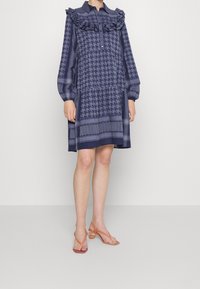 Granatowa sukienka z długimi rękawami w wzór houndstooth, z falbaniastego kołnierza i zapięciem na guziki. Noszona z pomarańczowymi sandałami na paski z okrągłym obcasem.