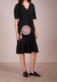 Bolso cruzado redondo de mimbre con círculos concéntricos en rosa, negro y blanco. Presenta un borlón y una correa trenzada. La modelo lleva un vestido negro.