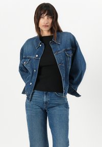 Veste en denim bleu foncé avec des garnitures en argent, fermeture par bouton et deux poches avant. Associée à un top noir en côte et à un jean bleu.