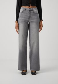 Personne portant un jean gris taille haute à jambes larges, un top noir court à manches longues et des ballerines noires cloutées, debout devant un fond uni.