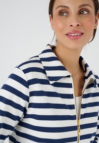 Giacca a righe in blu navy e bianco, con chiusura frontale a zip e colletto a punta, realizzata in tessuto testurizzato. Accento della zip dorata visibile.