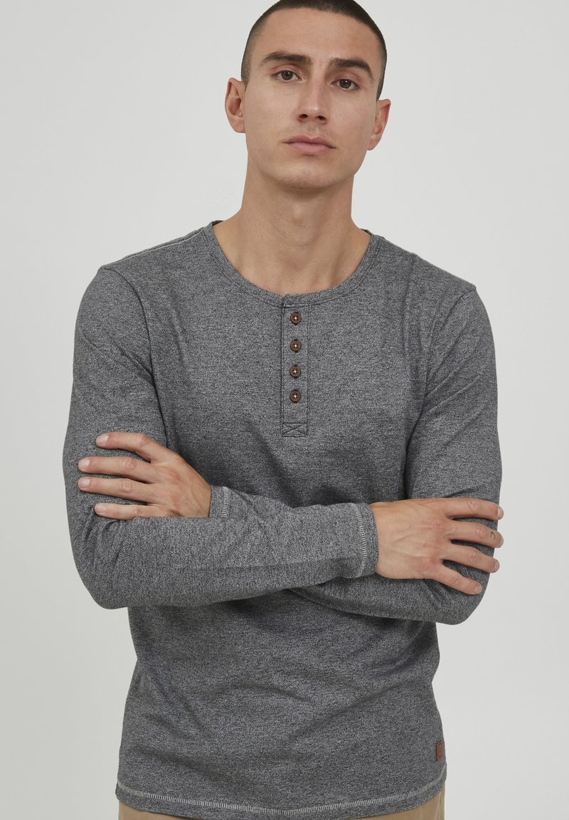 Chemise henley à manches longues grise avec un plastron à boutons, fabriquée en tissu texturé. Présente des coutures simples et une petite étiquette logo à l'ourlet.