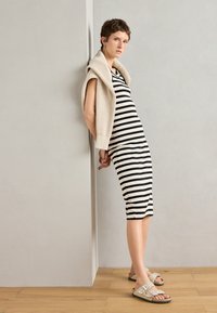 Robe midi à rayures noire et blanche associée à un cardigan en tricot beige. Présente des sandales plates avec des clous décoratifs. Silhouette moderne et ajustée.