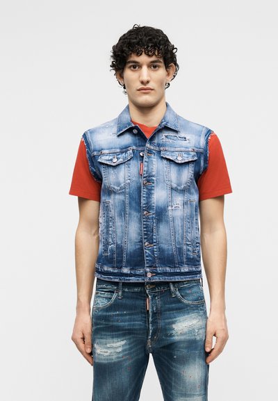 Νέος άνδρας με σγουρά σκούρα μαλλιά, φορώντας ένα φθαρμένο μπλε τζιν γιλέκο πάνω από ένα κόκκινο κοντομάνικο πουκάμισο και φθαρμένο μπλε τζιν παντελόνι.