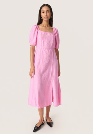 Mujer con vestido midi rosa con mangas abullonadas, escote cuadrado, abertura frontal y zapatos planos negros puntiagudos, de pie sobre un fondo sencillo.