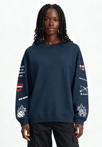 Donna con capelli intrecciati che indossa un felpa blu navy con simboli colorati, testo e stampe grafiche sulle maniche, abbinata a pantaloni neri.