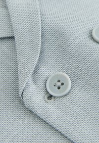 Närbild av texturerat ljusgrått tyg med två fastsydda fyrhålsknappar och överlappande vikta kanter.