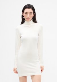 Rochie albă cu mânecă lungă și guler înalt, confecționată dintr-un material moale, cu o siluetă ajustată, având un accent cu un logo mic pe piept și manșete cu dunga.