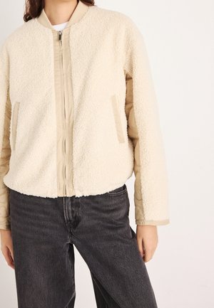 Kvinde iført en cremefarvet sherpa lynlåstrøje med beige quiltede ærmer og sorte jeans mod en hvid baggrund.