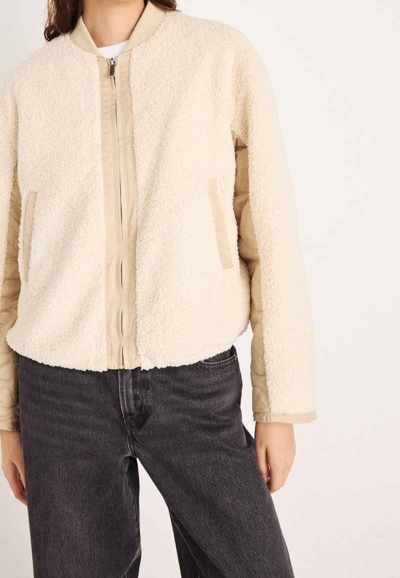 Femme portant une veste zippée sherpa crème avec des manches beige matelassées et un jean noir sur fond blanc.