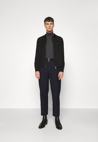 Veste noire avec deux poches poitrine, col roulé gris, pantalon bleu marine et bottes noires à la cheville, se tenant contre un fond blanc uni.