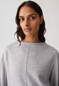 Donna con lunghi capelli scuri che indossa un maglione grigio chiaro a collo rotondo con una cucitura centrale, che guarda serenamente la camera su uno sfondo semplice.