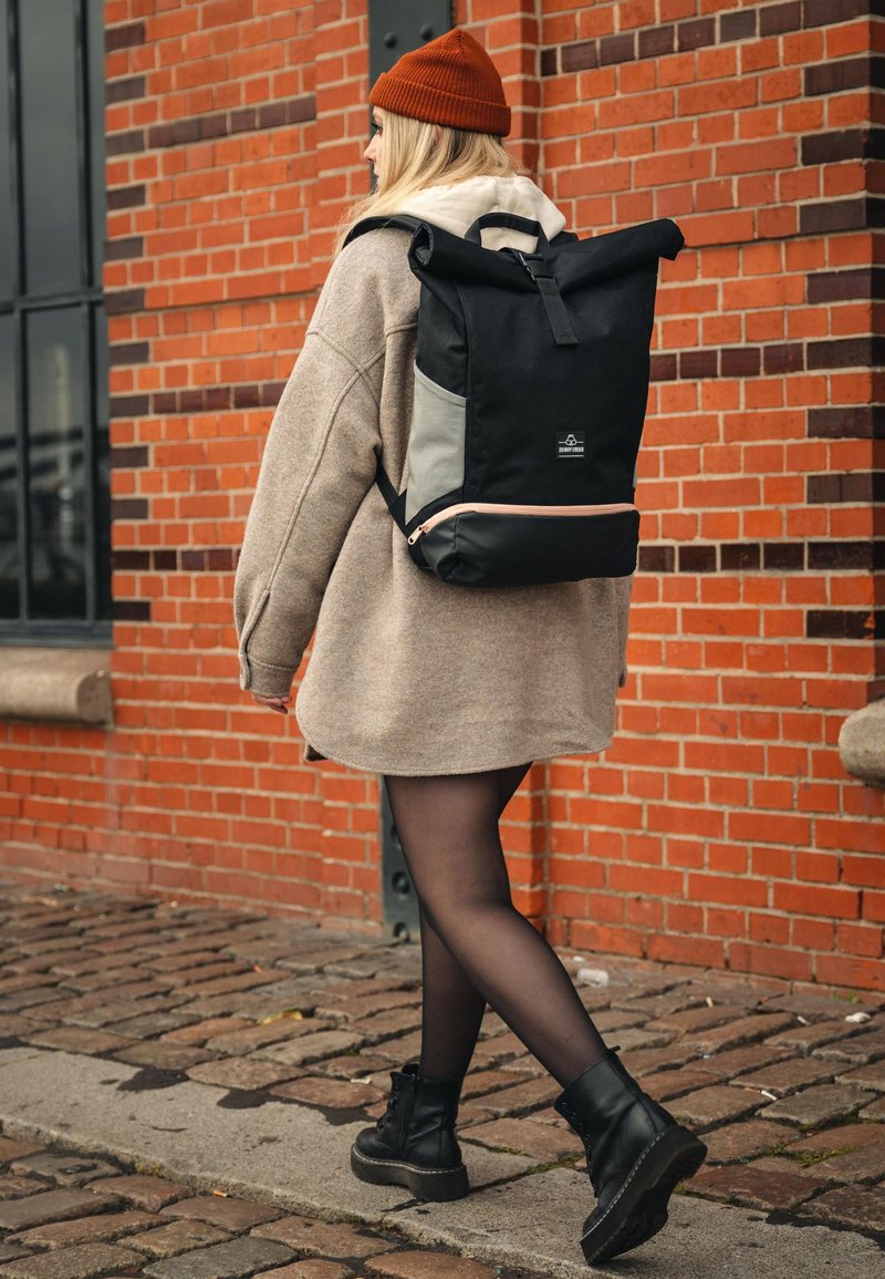 Ung kvinna i en beige kappa, svarta strumpbyxor och stövlar som går på kullerstenar bredvid en tegelvägg, bärande en svart och grå ryggsäck.