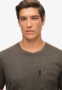 Ciemnooliwkowa bawełniana koszulka z okrągłym dekoltem, krótkimi rękawami i kieszonką na piersi z małą metką marki. Miękka tekstura, swobodny krój.