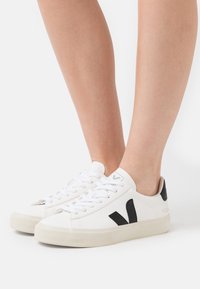 Weiße Sneakers mit schwarzen Akzenten, durchbrochenen seitlichen Details, runder Zehenform, flacher Sohle und Schnürverschluss. Logo auf Zunge und Ferse.