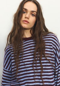 Prúžkovaný sveter v svetlomodrej a bordovej farbe. Mäkká textúra, široký okrúhly výstrih a voľný strih. Vlasy padajú vlnito, čo pridáva obrázku hĺbku.