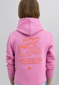 Roze hoodie, met een oranje geborduurd ontwerp van een koffiecup en croissant, met tekst die een café-adres en openingstijden detailleert.