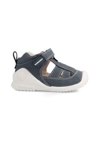 Grau genähten Schuh mit offenen Seiten, weißer Gummikappe, verstellbarem Riemen und strukturiertem Laufsohle. Markenlogo an der Seite sichtbar.