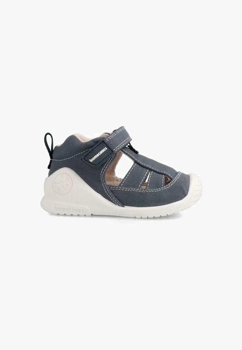 Sive obutev iz tkanine z odprtimi stranmi, bel gumijast zaščitni del na prstih, nastavljiv trak in teksturiran podplat. Logotip blagovne znamke vidno na strani.