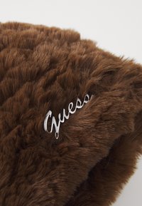 Brūna mākslīgā kažokāda ar mīkstu, fakturētu virsmu, pie sāniem piestiprināts sudraba metāla logotips ar uzrakstu "Guess".