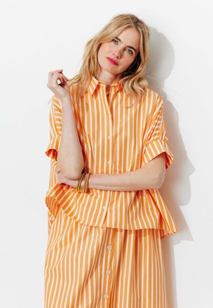 Femme blonde aux cheveux lâchés portant une chemise à manches courtes rayée verticale orange et blanche avec boutons et une jupe assortie, debout contre un mur blanc.