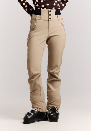 Vrouw die beige skibroek en zwarte skischoenen draagt, staat met handen in de zij tegen een effen achtergrond.