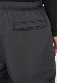 Pantaloni neri, leggeri e imbottiti, caratterizzati da una vita elastica e una tasca posteriore piatta con bordo cucito. Tessuto testurizzato e resistente all'acqua.